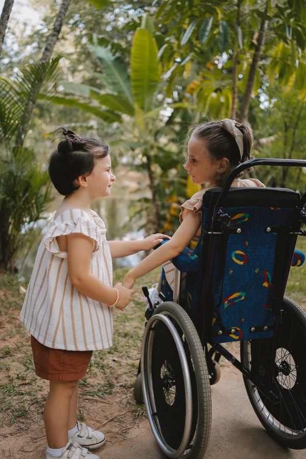 Duas meninas de mãos dadas em um jardim, uma delas em cadeira de rodas.